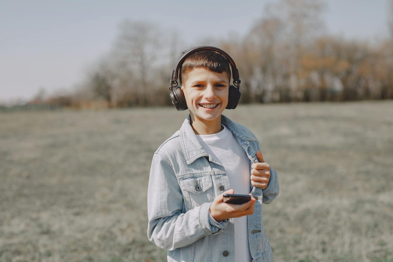 Garçon souriant portant un casque audio et une veste en jean en plein air au printemps.